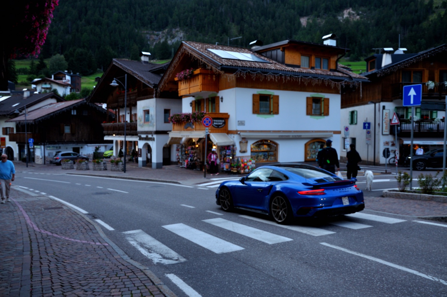 A blue Porsche crossing a zebra crossing in a mountain village at evening