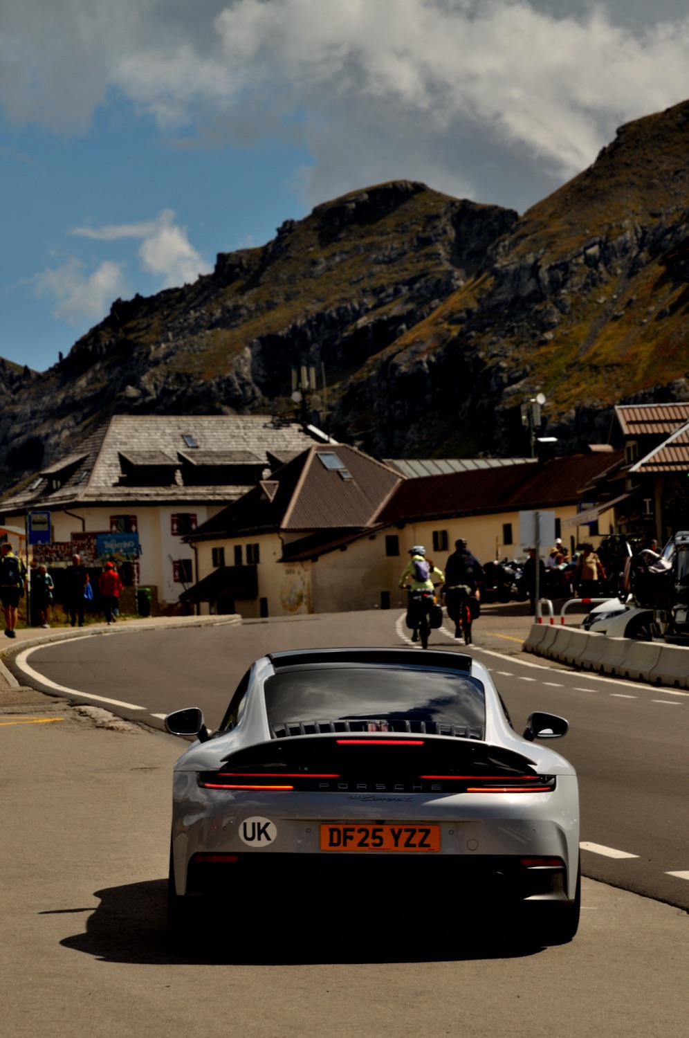 A silver Porsche 911 from behind on a mountain road, buildings and peaks ahead